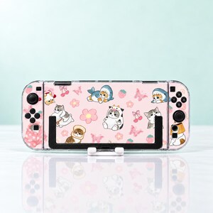 May include: A handheld gaming console featuring a light pink skin with a repeating pattern of cartoon cats in various costumes, including shark and cow outfits, alongside pink flowers, butterflies, cherries, and strawberries, held in a black stand.