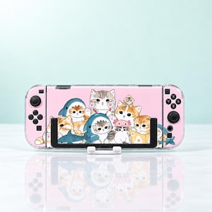 May include: A handheld gaming console featuring a light pink protective case decorated with a colourful illustration of multiple kittens, some wearing shark and axolotl costumes, resting on a white stand with a reflective surface below.