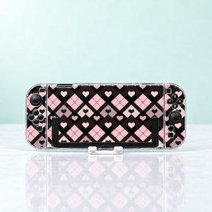 May include: A handheld gaming console featuring a protective skin with a black, pink, and white argyle pattern. The design includes small heart icons within the diamond shapes. The console has black joysticks, directional buttons, and action buttons.