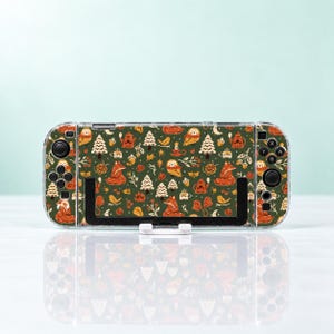 May include: A handheld gaming console featuring a dark green skin with a repeating pattern of orange foxes, white pine trees, brown owls, small mugs, and autumn leaves. The device has black buttons and joysticks, and is held in a black stand.