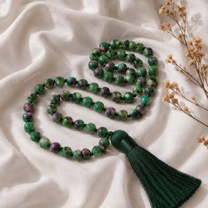 May include: A long necklace featuring round, polished beads of green zoisite with inclusions of deep magenta ruby. The beads are strung on a dark cord and terminate in a long, dark forest green silk tassel that hangs from a matching knotted cap.