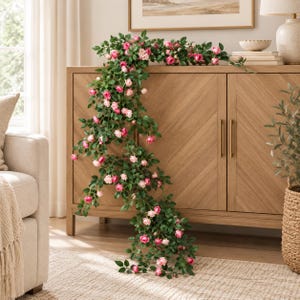 May include: A long, trailing artificial rose garland featuring numerous small, vibrant pink and soft pale pink flower blossoms interspersed with dense green foliage, cascading downwards from the top of a wooden cabinet surface.