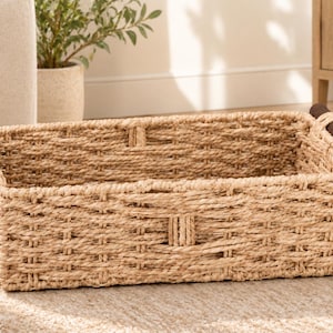 May include: A rectangular storage basket woven from light tan natural fibers, featuring a structured open-top design and two dark brown cylindrical wooden handles attached to the sides. The basket has a consistent, tight weave pattern throughout.