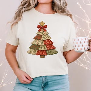 Könnte beinhalten: Ein cremefarbenes Kurzarm-T-Shirt mit einer Grafik eines Weihnachtsbaums im Patchwork-Stil. Der Baum besteht aus roten und grünen Stoffmustern mit Blumen- und Karodesigns und ist mit einem goldenen Stern und einer roten Schleife verziert.