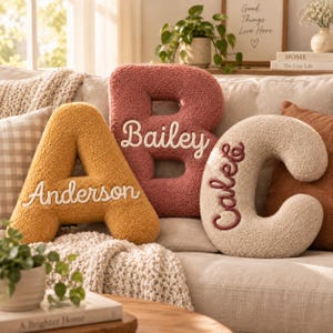 May include: Three plush, textured letter-shaped pillows arranged on a sofa. The yellow letter A features the white embroidered name Anderson, the terracotta letter B features the white name Bailey, and the cream letter C features the name Caleb.