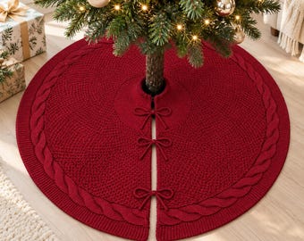 May include: A circular, deep red knitted Christmas tree skirt featuring a textured seed stitch pattern throughout the center and a thick, braided cable knit border along the outer edge. The skirt includes three matching red tie closures.
