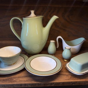 May include: A collection of sage green and white ceramic tableware featuring a tall teapot, a teacup with saucer, a small bowl, a gravy boat, a butter dish with lid, and two small shakers, all accented with thin gold trim along the edges.