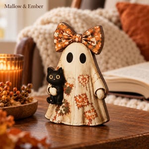 Pode incluir: Uma figura decorativa de fantasma em madeira com corpo cor de creme, padrões de colcha em laranja e castanho, um grande laço floral laranja no topo e um pequeno gato preto no braço. O texto Mallow & Ember é visível.