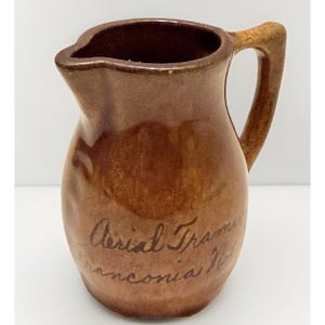 May include: A brown ceramic pitcher with a glossy finish and a curved handle. The front features dark handwritten cursive text that reads Aerial Tramway Franconia NH. The pitcher has a wide body, a narrow neck, and a distinct pouring spout.