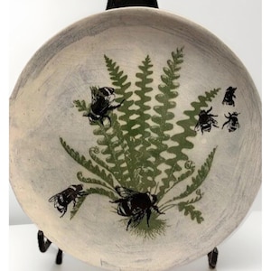 May include: A round ceramic plate with a light gray, textured surface featuring a central green fern illustration. Several black bumblebee silhouettes are positioned around the fern, with some bees appearing to land on the fronds of the plant.
