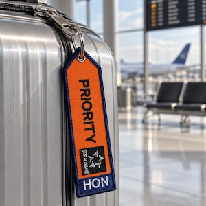 Op de afbeelding: Een rechthoekige oranje bagagelabel met een donkerblauwe rand bevestigd aan een zilveren koffer. Het label bevat het woord PRIORITY in het zwart, het Star Alliance-logo in een zwart vierkant en de letters HON in het wit op een blauwe achtergrond.