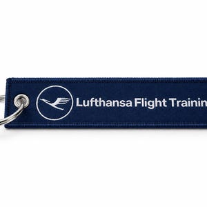 Könnte beinhalten: Ein rechteckiger Schlüsselanhänger aus dunkelblauem Stoff mit einem silbernen Metallring. Der Anhänger zeigt links das weiße Lufthansa-Kranich-Logo in einem Kreis und rechts den weißen Schriftzug Lufthansa Flight Training.