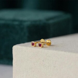 May include: A pair of gold stud earrings featuring square-shaped settings, each containing a single small, deep red gemstone. The earrings have a flat, circular backing and a post design, showcasing a polished metallic finish and vibrant red stones.