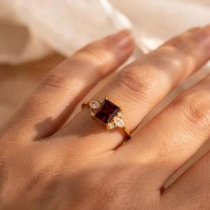 May include: A gold ring featuring a central rectangular deep red gemstone flanked by two smaller marquise-cut clear stones on each side. The band is thin and polished gold, showcasing a classic design with a vibrant, saturated red centre stone.