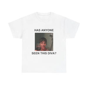 May include: A white short-sleeved t-shirt featuring a centered rectangular graphic of a person holding a glass of red wine in a shower setting. The text above the image reads HAS ANYONE and the text below the image reads SEEN THIS DIVA?