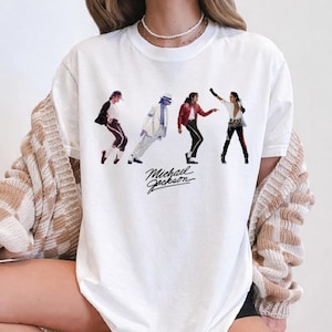 May include: A white crew neck t-shirt featuring four distinct graphic images of Michael Jackson in various iconic dance poses and outfits, with the name Michael Jackson printed in black cursive script lettering below the figures.
