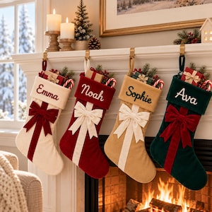 May include: Four velvet Christmas stockings hanging from a white mantelpiece. The stockings are cream, red, tan, and dark green, each featuring a large bow, a gift box, and a candy cane. They are embroidered with the names Emma, Noah, Sophie, and Ava.
