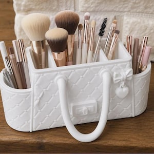 Puede incluir: Un organizador de brochas de maquillaje de plástico blanco con forma de bolso acolchado con dos asas, un lazo pequeño y un dije de corazón. El recipiente cuenta con múltiples compartimentos llenos de varias brochas de maquillaje y lápices cosméticos.