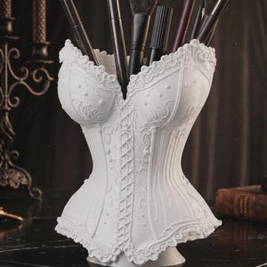 Peut inclure: Un organisateur de bureau blanc en forme de corset avec des motifs floraux en relief et un laçage central. L'intérieur creux contient plusieurs pinceaux de maquillage noirs, servant de récipient fonctionnel pour divers petits objets.