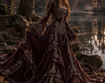 May include: A floor-length, deep burgundy velvet gown featuring intricate silver-toned embroidery, a skeletal ribcage design on the bodice, long flared sleeves, and a voluminous skirt with layered lace trim and cascading fabric panels.