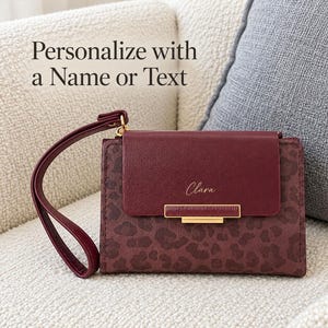 May include: A burgundy wristlet wallet featuring a dark leopard print pattern on the main body and a solid burgundy flap. The flap displays the name Clara in gold script and a gold metal bar accent. A matching wrist strap is attached.