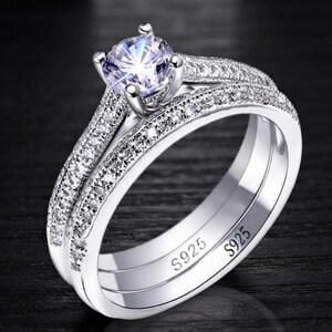 May include: A silver-toned ring set featuring a central round-cut clear stone held by four prongs. The band and the accompanying ring are encrusted with small clear stones along the top surface. The inner band is clearly stamped with S925.
