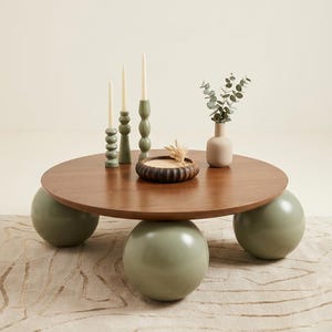 May include: A round wooden coffee table with a smooth brown top supported by three large, sage green spherical legs. On the table are three sage green candle holders with white taper candles, a small wooden bowl, and a beige vase with green leaves.