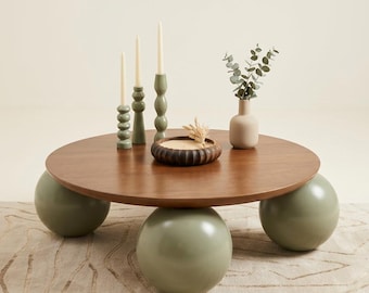 May include: A round wooden coffee table with a smooth brown top supported by three large, sage green spherical legs. On the table are three sage green candle holders with white taper candles, a small wooden bowl, and a beige vase with green leaves.