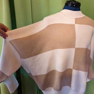 K&ouml;nnte beinhalten: Ein cremefarbener und beigefarbener Colorblock-Pullover mit Stehkragen. Der Pullover hat ein geometrisches Design mit abwechselnden horizontalen Streifen und quadratischen Mustern. Die &Auml;rmel sind lang und das Material scheint ein Strickstoff zu sein.