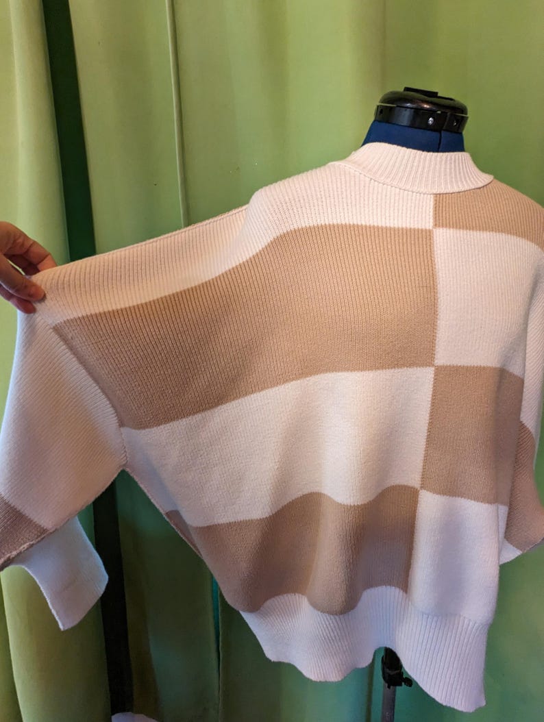 K&ouml;nnte beinhalten: Ein cremefarbener und beigefarbener Colorblock-Pullover mit Stehkragen. Der Pullover hat ein geometrisches Design mit abwechselnden horizontalen Streifen und quadratischen Mustern. Die &Auml;rmel sind lang und das Material scheint ein Strickstoff zu sein.