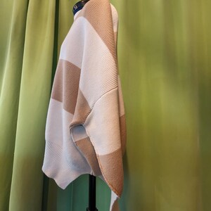 K&ouml;nnte beinhalten: Ein beige-cremefarbener gestreifter Pullover auf einer Schaufensterpuppe. Der Pullover hat breite Streifen und eine lockere Passform. Der Stoff scheint ein dicker Strick zu sein, der sich f&uuml;r k&auml;lteres Wetter eignet. Das Kleidungsst&uuml;ck ist drapiert und zeigt seine Form und sein Design.