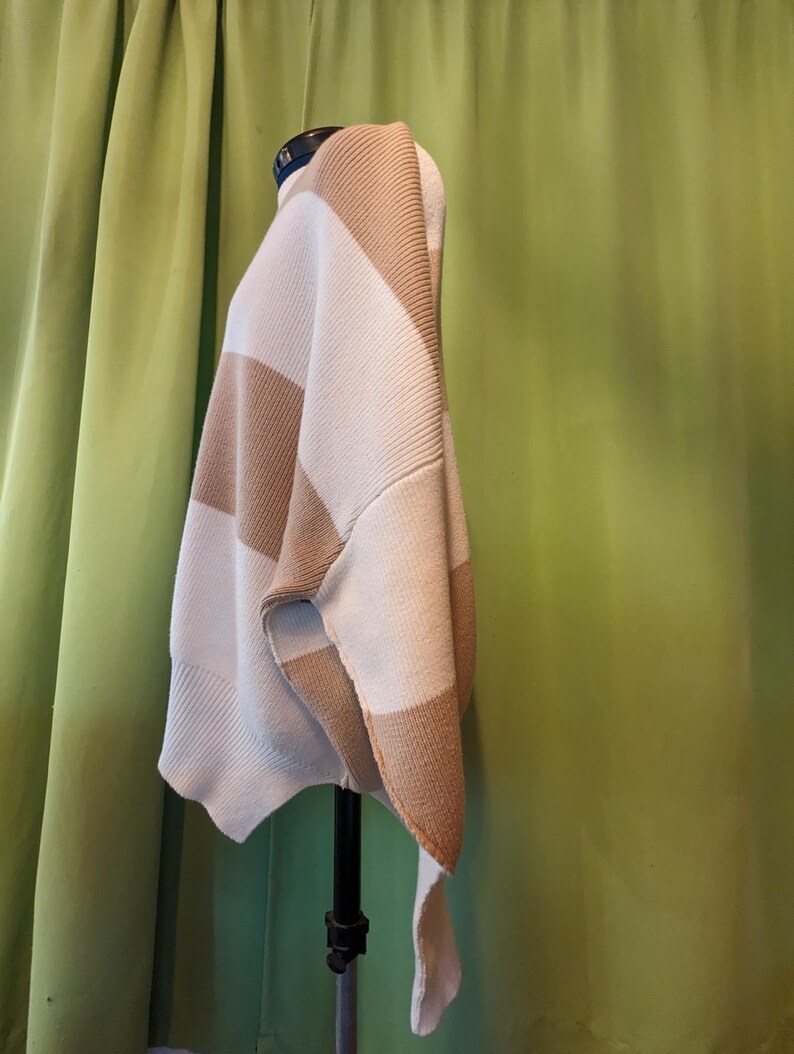 K&ouml;nnte beinhalten: Ein beige-cremefarbener gestreifter Pullover auf einer Schaufensterpuppe. Der Pullover hat breite Streifen und eine lockere Passform. Der Stoff scheint ein dicker Strick zu sein, der sich f&uuml;r k&auml;lteres Wetter eignet. Das Kleidungsst&uuml;ck ist drapiert und zeigt seine Form und sein Design.