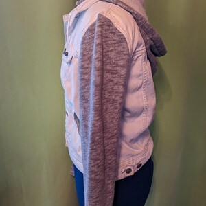 Op de afbeelding: Een lichtgekleurde spijkerjas met grijze gebreide mouwen en een bijpassende capuchon. De jas heeft knoopsluitingen en zakken. Het kledingstuk wordt op een mannequin getoond.