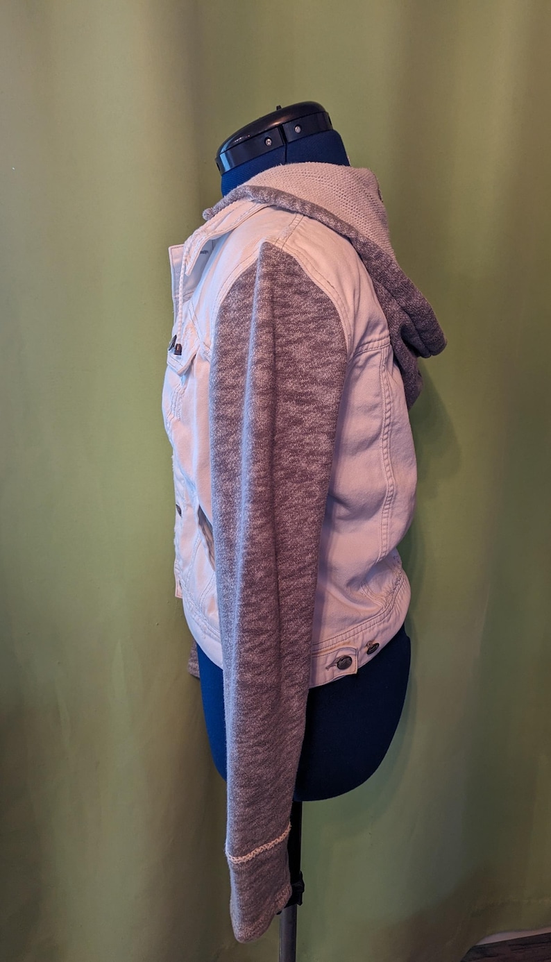 Op de afbeelding: Een lichtgekleurde spijkerjas met grijze gebreide mouwen en een bijpassende capuchon. De jas heeft knoopsluitingen en zakken. Het kledingstuk wordt op een mannequin getoond.