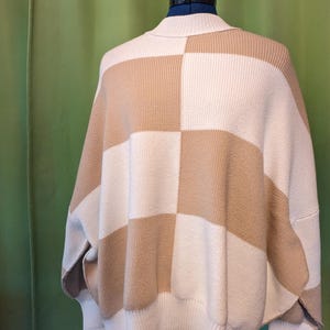 K&ouml;nnte beinhalten: Ein beige-wei&szlig; karierter Strickpullover mit weitem, entspanntem Schnitt. Der Pullover hat einen hohen Kragen und eine einzigartige, kastenf&ouml;rmige Silhouette. Das Farbschema ist neutral, mit abwechselnden Quadraten in Beige und Wei&szlig;.
