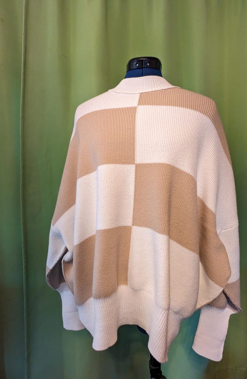 K&ouml;nnte beinhalten: Ein beige-wei&szlig; karierter Strickpullover mit weitem, entspanntem Schnitt. Der Pullover hat einen hohen Kragen und eine einzigartige, kastenf&ouml;rmige Silhouette. Das Farbschema ist neutral, mit abwechselnden Quadraten in Beige und Wei&szlig;.