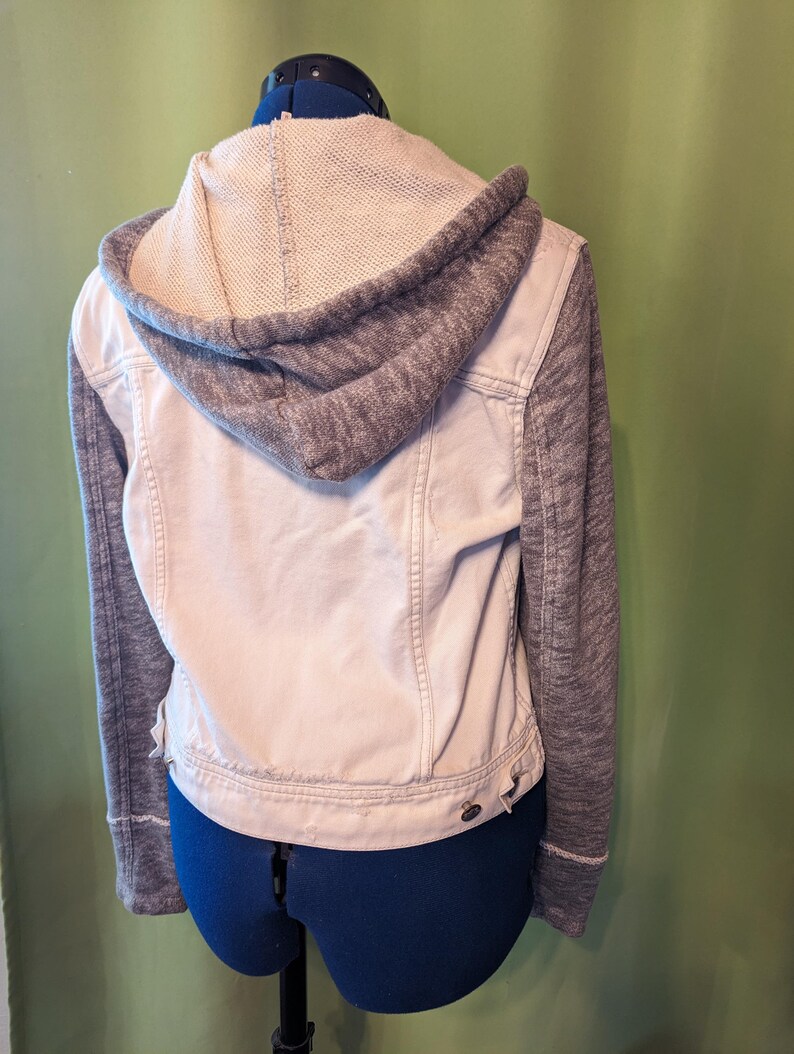 Op de afbeelding: Een lichtbeige spijkerjasje met een grijze gebreide capuchon en mouwen. De jas heeft een knoopsluiting en een klassiek ontwerp, perfect voor casual wear. De achterkant van de jas is zichtbaar en toont de stikseldetails.