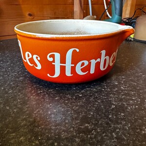 May include: A round orange ceramic mixing bowl with a white interior and a small pouring spout. The exterior features the white text "les Herbes" in a decorative script font. The bowl has a smooth finish and a wide, open top design.
