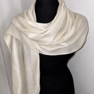Peut inclure: Une écharpe unie blanc cassé avec une finition lisse et lustrée drapée autour d'un mannequin noir. Le tissu présente un éclat subtil et des plis souples, créant un aspect superposé autour du cou avec une extrémité tombant sur le côté.