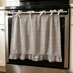 Puede incluir: Una cortina para horno con estampado de cuadros vichy en marrón y blanco, que presenta un dobladillo con volantes en la parte inferior y cinco lazos de tela sujetos a la barra superior. La cortina es de tela tejida con un patrón uniforme.