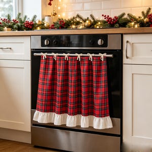 May include: A red, green, and black plaid oven towel hanging from a stainless steel oven handle. The towel features six small white fabric bows along the top edge for attachment and a white ruffled lace trim along the bottom hem.