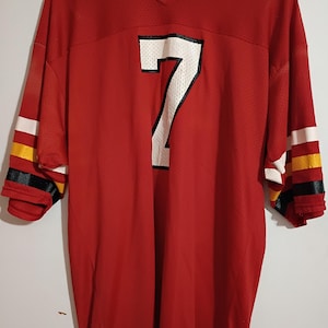 May include: A red mesh V-neck jersey featuring the number 7 in white with a black outline on the chest. The sleeves have horizontal stripes in white, yellow, and black. A Sand-Knit brand tag is visible on the lower front hem of the garment.