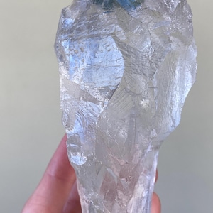 May include: A large, raw, translucent clear quartz crystal specimen with a jagged, irregular shape. The surface features numerous natural facets, sharp edges, and internal fractures that catch the light, creating a reflective, glassy appearance.