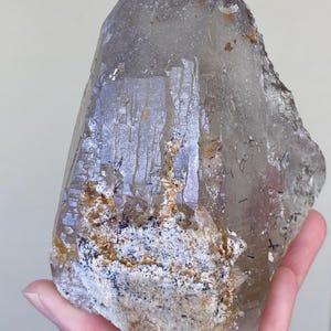 May include: A large, translucent smoky quartz crystal point featuring a jagged, textured base with inclusions of white mineral deposits, golden-brown iron staining, and thin, needle-like black tourmaline crystals embedded throughout the structure.
