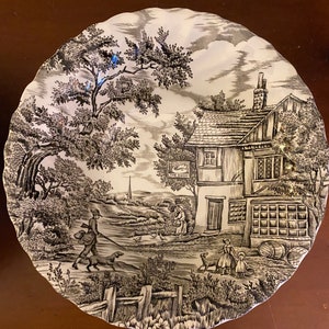 Puede incluir: Un plato decorativo en blanco y negro con una escena de paisaje rural. El plato presenta una ilustración detallada de un pueblo con personas, perros y un edificio con el letrero "The Swan". El plato tiene un borde festoneado.