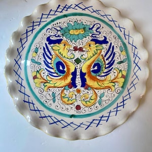 May include: A decorative ceramic plate with a scalloped edge. The plate features a central design of two stylized birds in yellow, orange, and blue, surrounded by floral and scrollwork patterns. The rim has a blue crosshatch design and a green border.