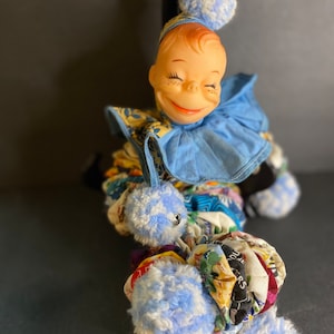 Puede incluir: Un payaso de trapo vintage con un traje de tela azul y blanco. El payaso tiene una cara sonriente con los ojos cerrados y un sombrero de pompón rojo.