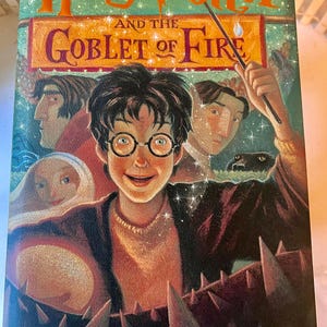 May include: A book cover for "Harry Potter and the Goblet of Fire" by J.K. Rowling. The cover features a smiling Harry Potter with round glasses, surrounded by other characters and a dark background. The title is in orange letters.