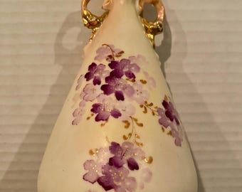 May include: Cream ceramic vase with a flared top and two ornate handles. The vase is decorated with purple floral designs and gold accents. The vase has a bulbous shape and is approximately 18 cm tall.