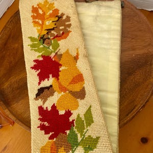 May include: A long, rectangular needlepoint wall hanging featuring a cream-colored background with a vertical pattern of colorful autumn leaves in shades of red, orange, yellow, and green, finished with a metal hanging bar and a circular ring.